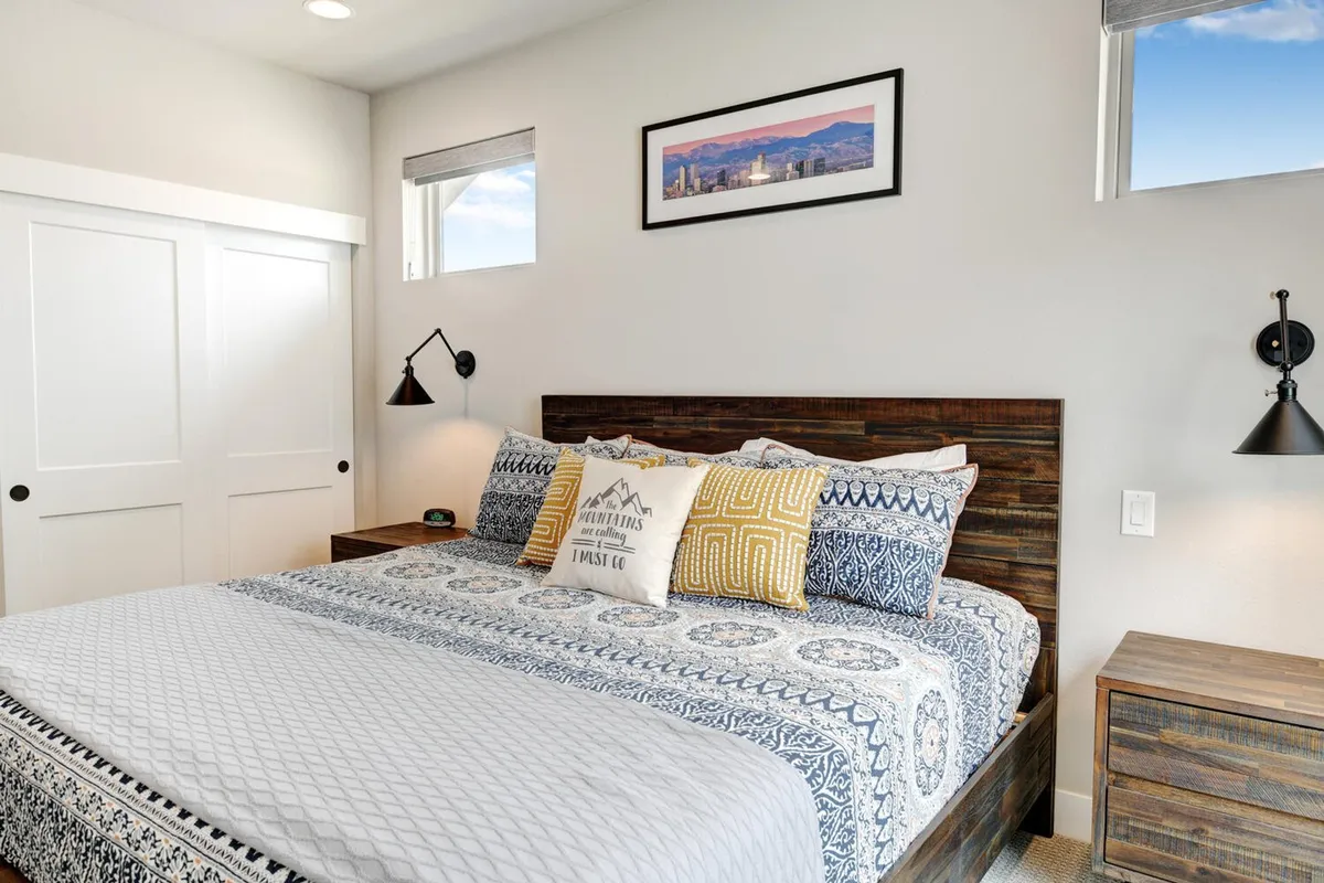 A beautifully styled bedroom centered around a plush bed with patterned bedding, paired with tasteful wall art and matching nightstands topped with lamps. The atmosphere is warm and inviting, with natural light suggesting the presence of blackout shades for restful sleep.