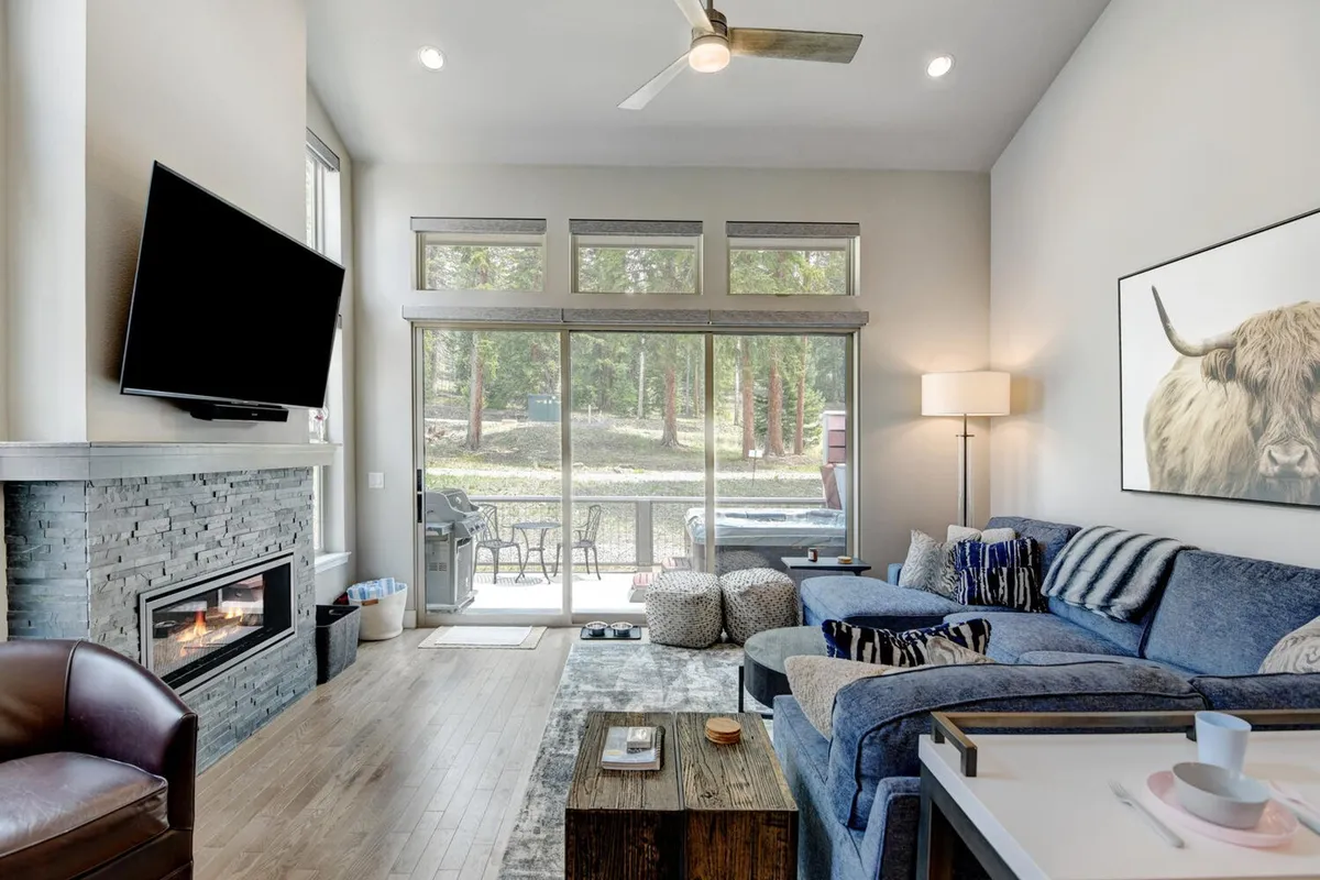 A spacious living area featuring a blue sectional sofa arranged around a sleek modern fireplace and a wall-mounted HD TV. Large windows provide views of surrounding trees and let in abundant natural light, while unique mountain-inspired artwork adds charm. The space is styled for cozy gatherings and relaxation.