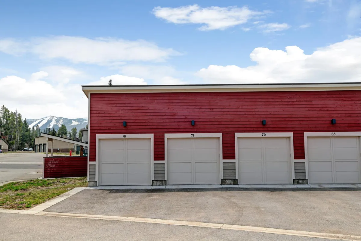 The home's red wooden garage facade stands against a majestic mountain backdrop, signaling both practical amenities and easy access to mountain adventures. The setting suggests space for gear and transitions from home life to the outdoors.