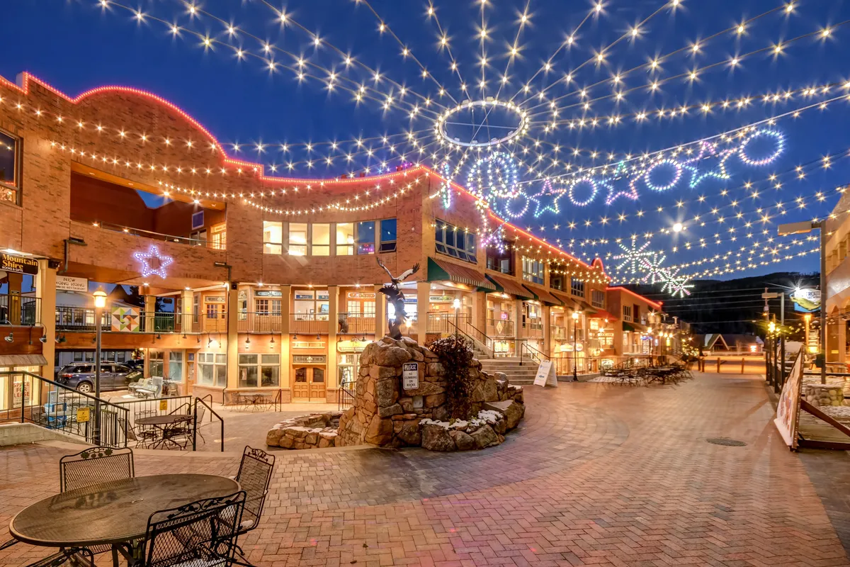 The image features string lights glowing above café tables, decorative stars, a stone fountain, and pedestrian spaces—evoking the festive atmosphere of downtown Winter Park’s outdoor dining and socializing spots. The joyful mood and evening lighting reflect community events and seasonal gatherings.