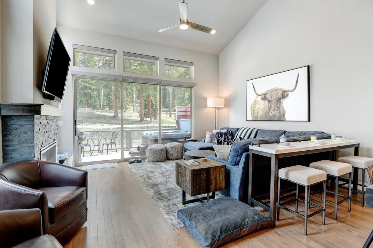 A cozy living room scene featuring a comfortable sofa, artwork of a cow above the fireplace, large windows revealing a forested landscape, and bar stools along a kitchen counter. The space is bathed in natural light, suggesting a strong connection to the outdoors.