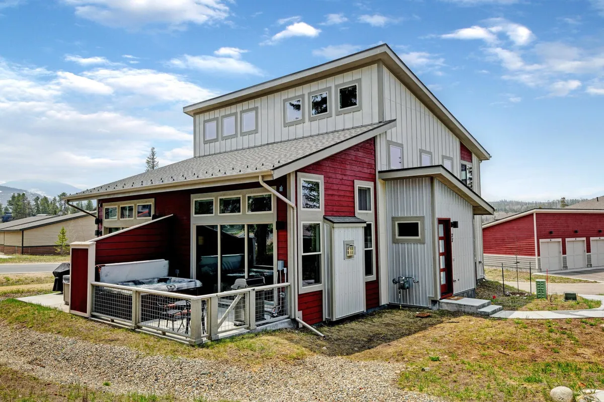 The hero image showcases the exterior of a modern mountain cabin in Fraser, Colorado, featuring striking red and white architecture, expansive windows that capture mountain views, and a spacious patio set for outdoor dining and relaxation. The scene is bathed in a warm sunset glow, inviting guests to envision evenings spent with friends and family outdoors.