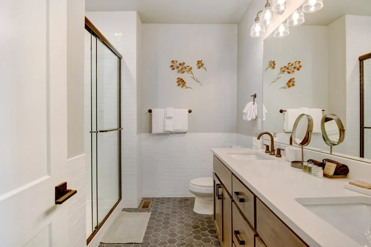 A modern, spa-inspired bathroom with a spacious shower enclosure, double sink vanity, floral wall decor, and stylish hexagonal tile flooring. The space gleams with natural light, enhancing the sense of luxury and relaxation.