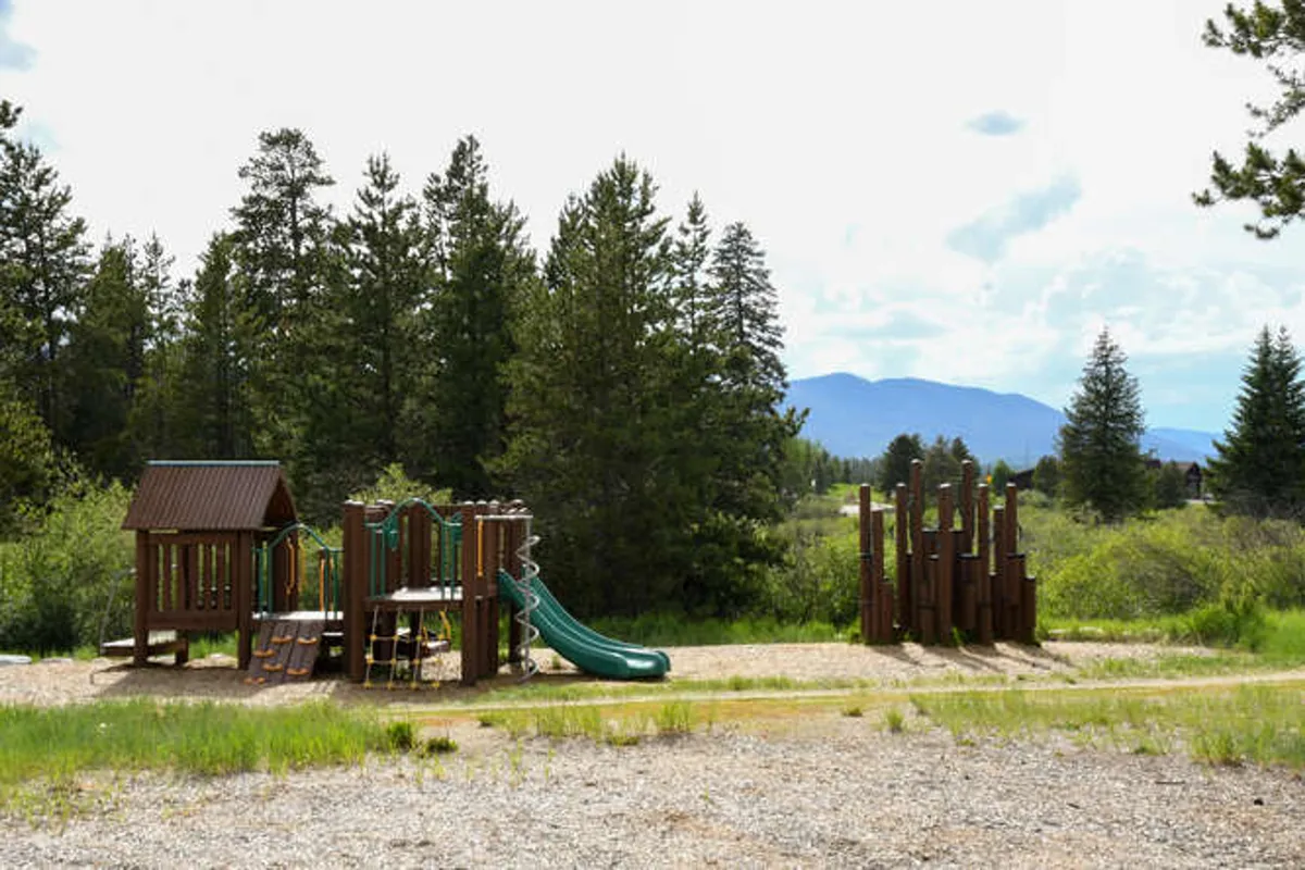 An outdoor playground set amidst trees and with a mountain backdrop, showing green slides and space for children to play. The scene is joyful, spacious, and perfect for families seeking kid-friendly amenities and outdoor fun just steps from the home.