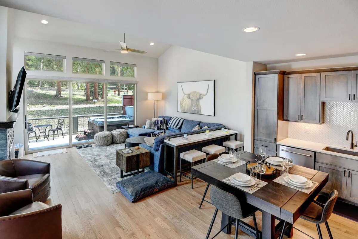 An open-concept area that combines a comfortable seating zone with a dining table set for a meal. Expansive windows flood the space with morning light and offer serene views of trees, while decorative art enhances the inviting ambiance. The arrangement encourages socializing and togetherness.